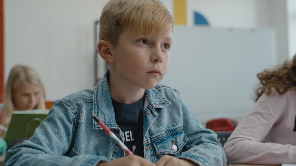 A young boy in a denim jacket attentively learning in a classroom setting.