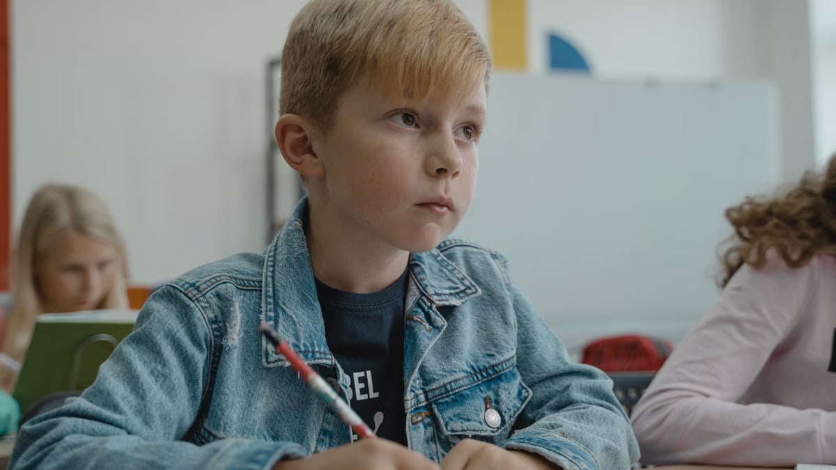 A young boy in a denim jacket attentively learning in a classroom setting.